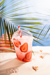 Cold fresh pink cocktail with ice in plastic mug, sun hat on tropical beach, vertical. Vacation and picnic summer on ocean coast.