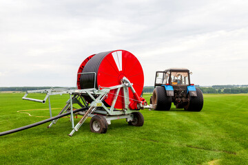 Watering a vegetable green field with a big water hose on the red bobbin near blue tractor in summer. Agriculture and food producing.