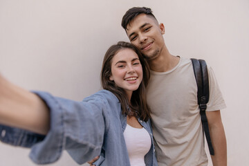 Selfie-portrait of young couple isolated over light background. Pretty dark-haired girl with pure skin, white top and blue shirt posing with attractive boyfriend, wearing beige t-shirt