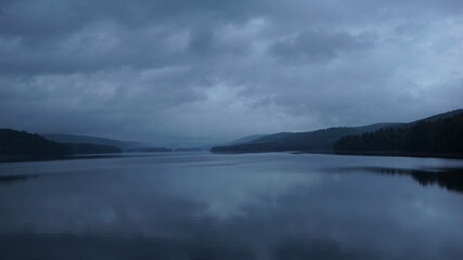 Clouds hover over a still lake on a grey evening.  Mountain background.