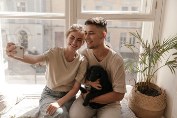 Pretty young couple with black dog making selfie. Adorable lady in loungewear sitting on windowsill, smiling and holding phone near husband, against window background - Powered by Adobe