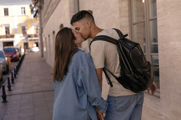 Lifestyle portrait of young couple walking on street and kissing. Cute dark-haired boy wearing beige t-shirt and black backpack posing with pretty girl in denim shirt