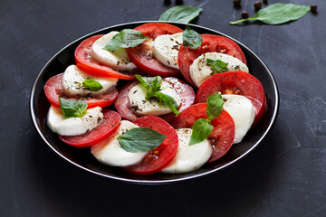 close-up traditional Italian caprese salad with green and spice on the black plate with copy space. sliced red tomatoes, mozzarella cheese, green basil, olive oil on a dark background. fresh salad.