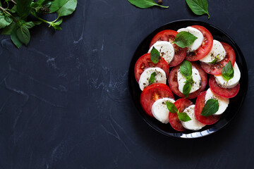 traditional Italian caprese salad with green on the black plate with copy space. sliced red tomatoes, mozzarella cheese, green basil, olive oil on a dark background. fresh salad.top view.