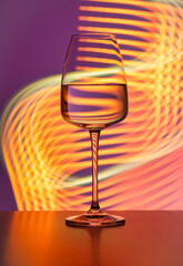 Crystal glass with pure water on a purple background painted with colorful light. No splashes, even surface. Light reflections and refractions, light streaks, party atmosphere, lifestyle, abstract.