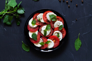 traditional Italian caprese salad with green and spice on the black plate with copy space. sliced red tomatoes, mozzarella cheese, green basil, olive oil on a dark background. fresh salad.top view.
