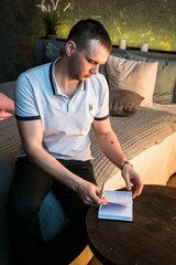 A young man is leafing through a notebook on a wooden table. A man is sitting on a bed in a loft-style room. Stylish interior.