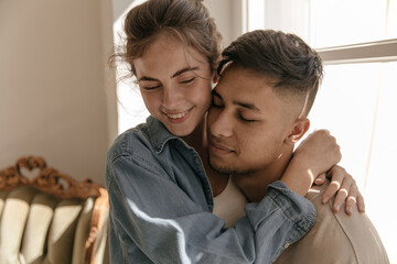 Lifestyle portrait of young couple hugging in sunlit room. Pretty dark-haired lady in blue shirt...