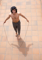 Young man doing skipping exercise barefoot on terrace outdoor with cap and headphones smiling
