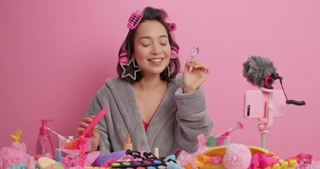 Happy beautiful Asian woman applies mascara does makeup and hairstyle records broadcasting video dressed in soft bathrobe isolated over pink background shows product to audience. Vlogging concept