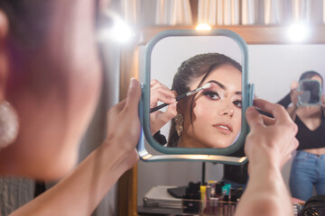 Beautiful young girl holding a mirror while the makeup artist smudges shadows on her eyes.