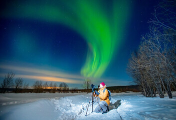 Photographer man with camera and tripod photographs aurora borealis, northern lights green. Concept photo tour to arctic travel - obrazy, fototapety, plakaty