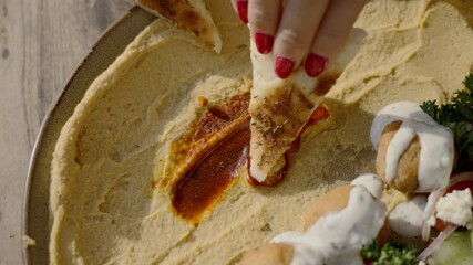 Female hand holding a piece of pita bread dipping it into hummus and paprika sauce. Falafel and...