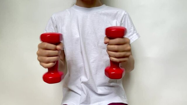 A girl in a white T-shirt slowly moves two red dumbbells weighing 1 kg 