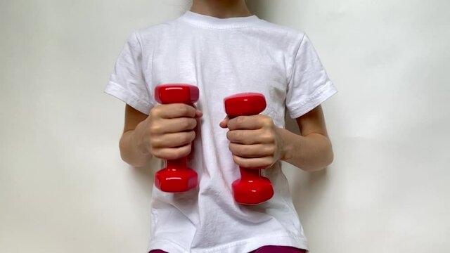 A girl in a white T-shirt slowly moves two red dumbbells weighing 1 kg 