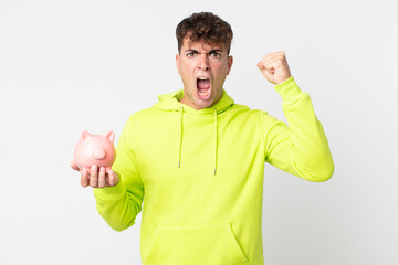 young handsome man shouting aggressively with an angry expression and holding a piggy bank
