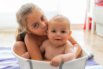 Closeup of Caucasian siblings together in a portable bathtub. Girl kisses baby boy.
