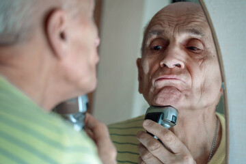 Elder grey haired man looks in mirror and holds electrical shaver near chin