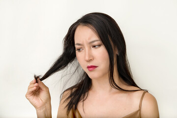 a young attractive Asian woman looks with displeasure at the tips of dry hair on a white background, the problem is the cross-section of the ends on the hair. the concept of hair care, hair nutrition.