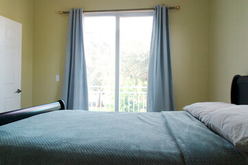 Full size bed fills room in front of sunlit window door leading to baclony