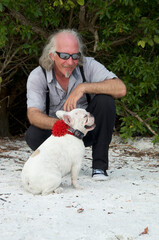 A handsome smiling long haired man with sunglasses is at beach petting small dog