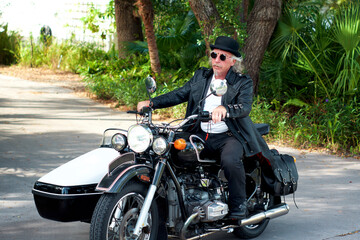 A handsome blond haired, goatee wearing steampunk biker driving black and white sidecar motorbike. Wearing leather tailcoat, bowler hat and round sunglasses with hair blowing in breeze