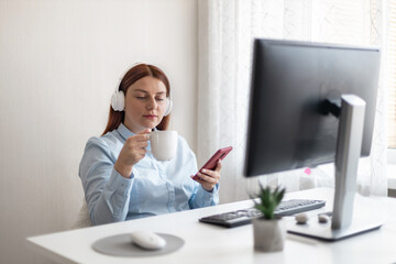 Obraz premium Young caucasian office woman worker sitting in wireless headphones with a cup of hot coffee or tea at the workplace at home or in the office. Lunch break. Using a smartphone