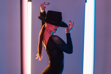 Alluring woman in hat between flashing colored neon lights, studio background. Femme fatale, stylish outfit of ballet dancer.