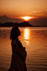 Pregnant woman in profile at sunset. Side view of silhouette in rays of setting sun reflected in water of beautiful woman standing on seashore. 