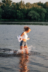 Photo of a running blonde in profile. A young woman in a white top and shirt and denim shorts runs freedom on the water against the background of a beautiful landscape at sunset. 