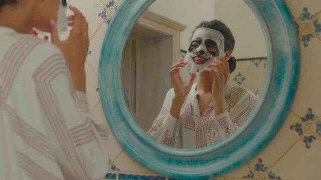 Close up of young adult woman applying facial cosmetics mask looking like panda. Mirror reflection of woman in panda mask.