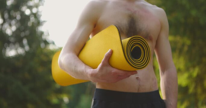 Close-up shot of Portrait of Young Man Yog. He Standing in Park with Sport Mat in Hands and Looking forward. Yoga Training in Park. Concept of Healthy Lifestyle. Slow motion