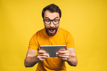 Amazed happy bearded man using digital tablet looking shocked about social media news, astonished man shopper consumer surprised excited by online win isolated over yellow background. - obrazy, fototapety, plakaty