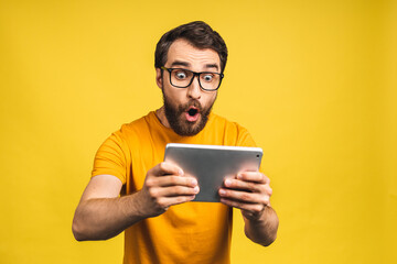 Amazed happy bearded man using digital tablet looking shocked about social media news, astonished man shopper consumer surprised excited by online win isolated over yellow background. - obrazy, fototapety, plakaty