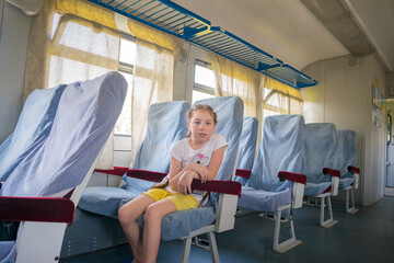 The child sits in an empty electric train carriage. A little girl sits on herself in the train compartment. A small passenger of railway transport.