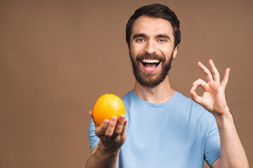 Healthy food and diet concept. Portrait of young bearded smiling man holding an orange fruit isolated on beige background.