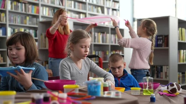 Cheerful female teacher and girl inflating slime bubble, boy with cerebral palsy looking at sister playing slime sitting next. Children with different disabilities during inclusive masterclass