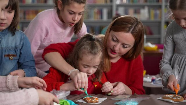 Close-up of teacher helping little girl with autism to decorate biscuit with glaze. Kids with disabilities decorating cookies with colorful icing during masterclass