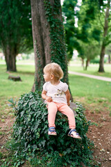 Little girl sits on a tree stump entwined with green ivy in the park, turning her head