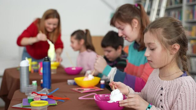 Two girls with psychoorganic syndrome enthusiastically making slimes on foreground. Boy with autism doing nothing while female teacher helping excited girl with cerebral palsy on background