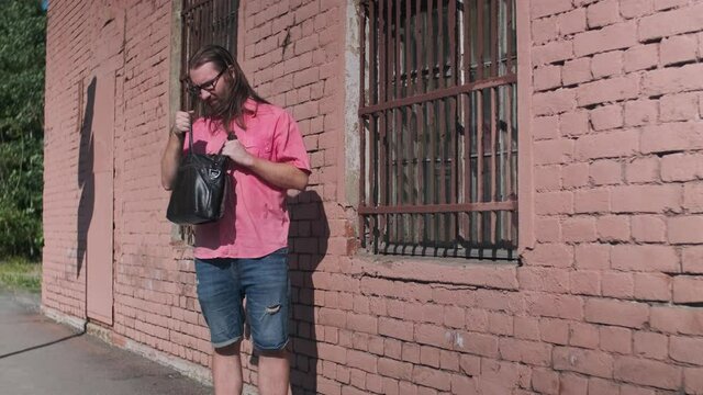 A man seems to be a drug dealer. person who just lost something in his handbag and looking for it. long haired male wearing glasses is waiting for someone near a painted in dead salmon brick building