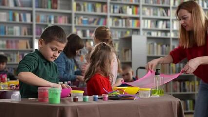 Joyful female teacher and little girl with autism stretching slime to inflate bubble during masterclass for kids with disabilities. Healthy children and kids with special needs spending time together - Powered by Adobe
