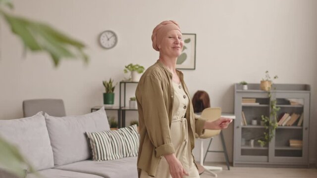 Medium shot of cheerful mature woman in headscarf celebrating cancer remission dancing alone in living room