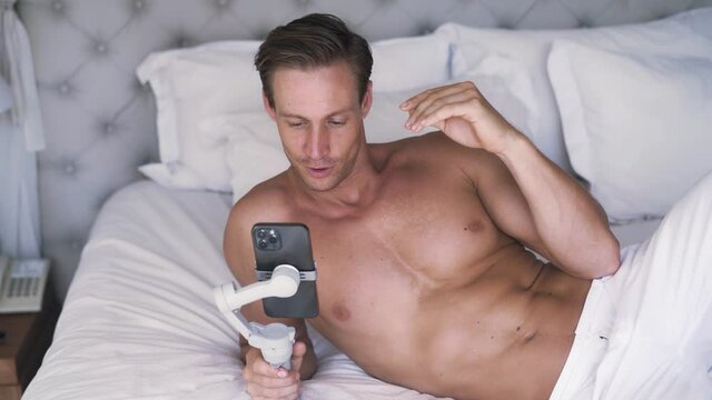 Portrait of young Caucasian man have a video call on his smartphone. He's laying on white sheets, have a video call and wave goodbye. Close up portrait lifestyle. High quality 4k footage video.