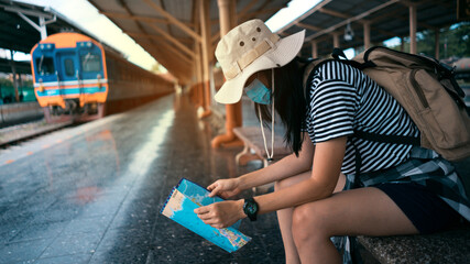 Fototapeta na wymiar New normal traveler concepts. Young female tourist wearing a mask looks at a tourist map while waiting for a train at the train station.