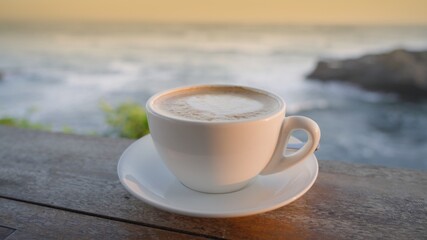 A beautiful still life with a cup of coffee latte on a wooden table of beach bar with ocean waves in the golden hour light on background. Enjoy summer vacation at seaside. Tasty cappuccino on a beach.