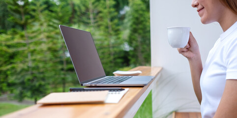 Closeup image of a young asian woman drinking coffee while using and working on laptop computer in the outdoors