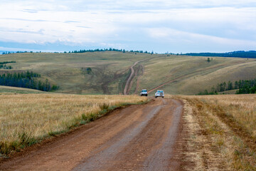 Two off-road cars are driving along the hills of steppe on dirt road. Horizontal image.