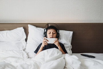 Serious asian boy in headphone lying on the bed while playing game on a smart phone