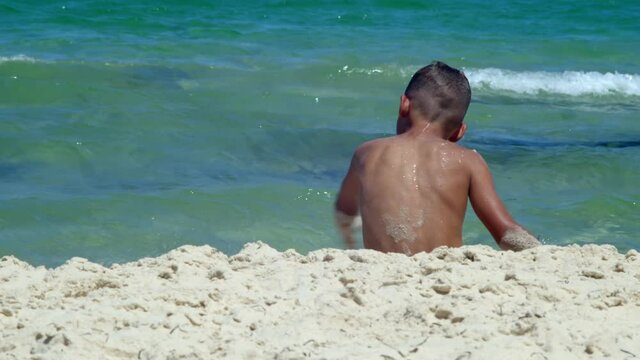 A tanned boy is sitting on the white sand by the sea with his back to the camera and playing with the sand. holidays at sea resorts with children. happy childhood by the sea. games by the sea under
